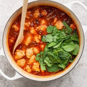 A white pot filled with tomato-based stew, meatballs, and fresh spinach, stirred with a wooden spoon on a light textured surface.