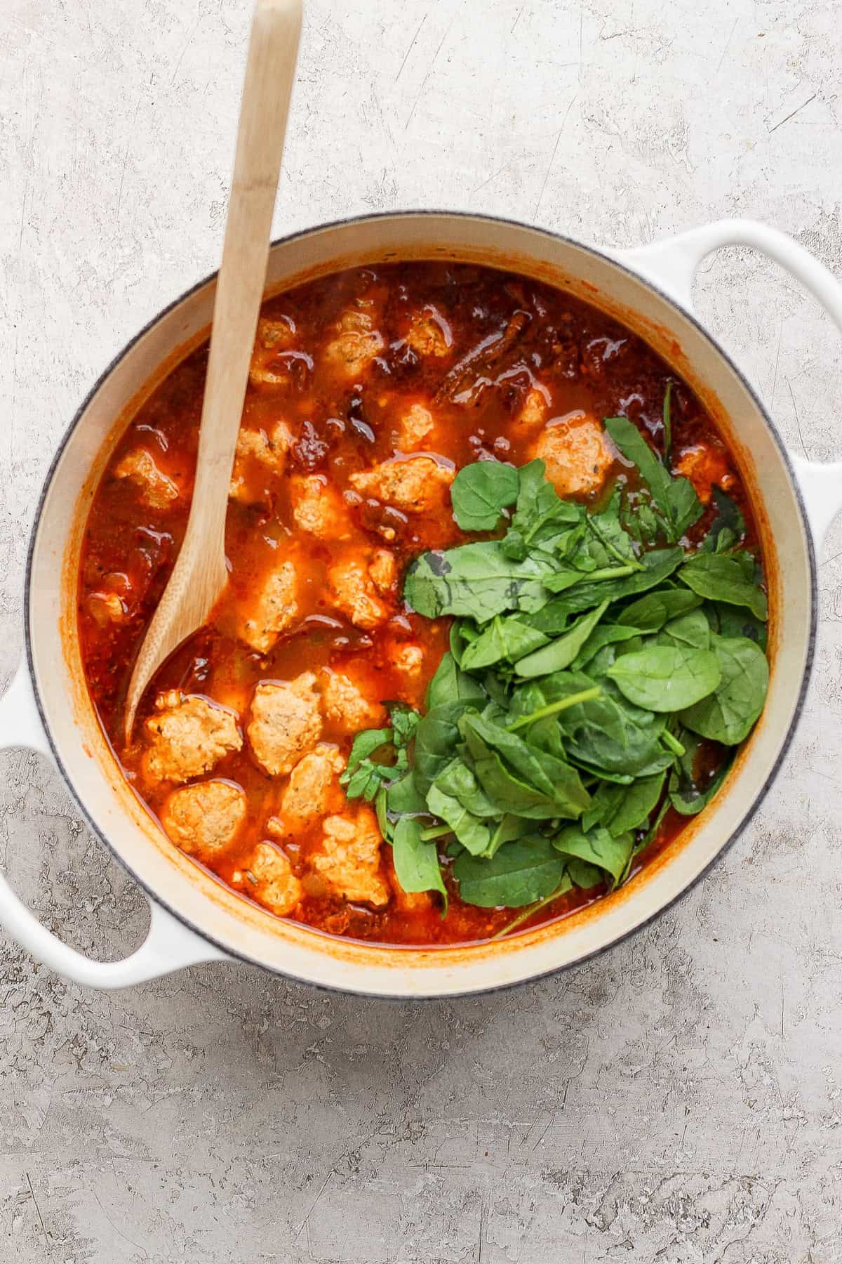 A white pot filled with tomato-based stew, meatballs, and fresh spinach, stirred with a wooden spoon on a light textured surface.