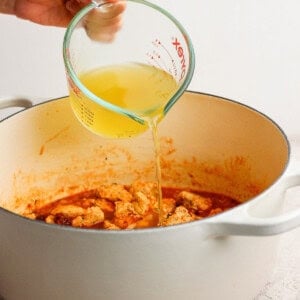 A hand pours chicken broth from a glass measuring cup into a white pot filled with cooked meat and sauce.