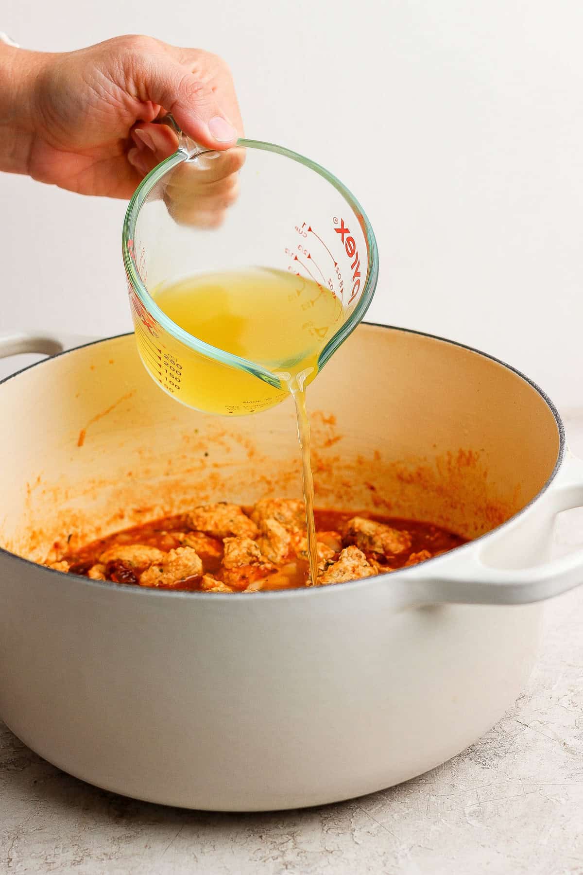 A hand pours chicken broth from a glass measuring cup into a white pot filled with cooked meat and sauce.