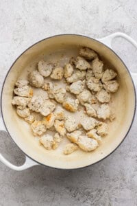 Chunks of cooked chicken are scattered in a white enameled pot on a light textured surface.