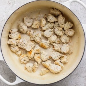 Chunks of cooked chicken are scattered in a white enameled pot on a light textured surface.