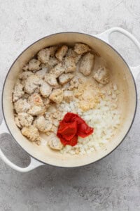 A white pot contains cooked chunks of sausage, chopped onions, minced garlic, and tomato paste on a light countertop.