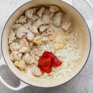 A white pot contains cooked chunks of sausage, chopped onions, minced garlic, and tomato paste on a light countertop.