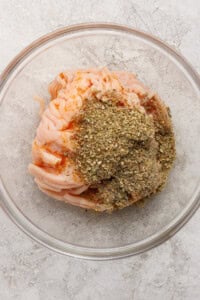 Raw ground meat topped with dried herbs and spices in a clear glass bowl on a light textured surface.