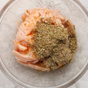 Raw ground meat topped with dried herbs and spices in a clear glass bowl on a light textured surface.