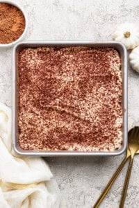 A square pan of tiramisu topped with cocoa powder sits on a light textured surface next to a bowl of cocoa, white cloth, gold fork and spoon, and two small white pumpkins.