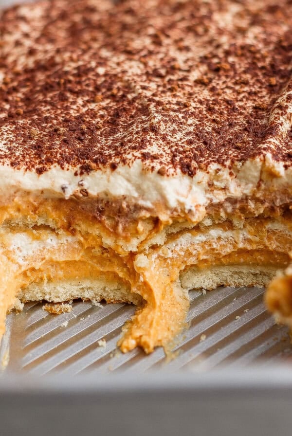 A metal baking pan with a partially sliced layered dessert featuring cream, a pumpkin-colored layer, and cocoa powder sprinkled on top.