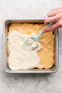 A hand uses a blue spatula to spread a white creamy layer over a yellow batter in a square metal baking pan.