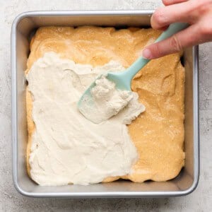 A hand uses a blue spatula to spread a white creamy layer over a yellow batter in a square metal baking pan.
