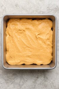 A square metal baking pan filled with an even layer of light brown cake batter on a textured light surface.