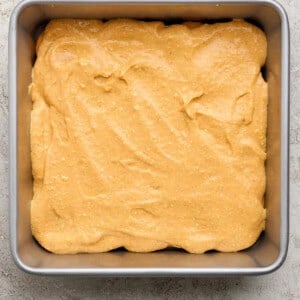 A square metal baking pan filled with an even layer of light brown cake batter on a textured light surface.