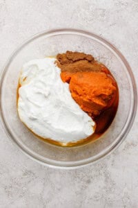 A glass bowl containing pumpkin puree, Greek yogurt, maple syrup, and spices on a light textured surface.