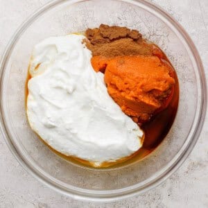 A glass bowl containing pumpkin puree, Greek yogurt, maple syrup, and spices on a light textured surface.