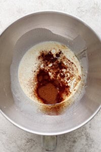 A metal mixing bowl contains a mixture of cream and sprinkled ground spices, including cinnamon, on a light textured surface.