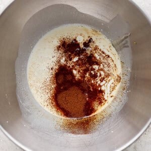 A metal mixing bowl contains a mixture of cream and sprinkled ground spices, including cinnamon, on a light textured surface.